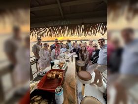 A lively group gathers for a sunset celebration on a boat, enjoying food and drinks while taking in the beautiful view — perfect for memorable gatherings and special occasions.