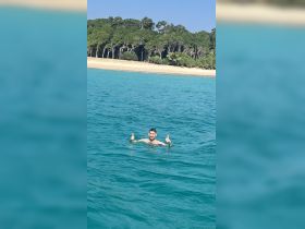A man enjoys a refreshing swim in crystal-clear waters, giving a thumbs up while surrounded by a beautiful beach backdrop — perfect for summer adventures.