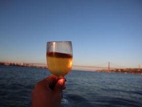 A hand holds a refreshing cocktail against the backdrop of a stunning sunset and a bridge, perfect for enjoying drinks on a boat trip.