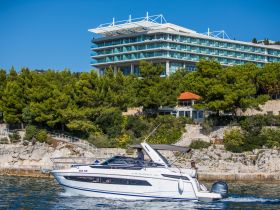 A sleek yacht is docked near a coastal hotel, showcasing its elegant design against a backdrop of lush greenery and clear blue skies.