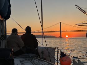 A couple enjoys a breathtaking sunset view from their sailboat, with a stunning bridge silhouette in the background — a perfect moment for romantic sailing experiences.