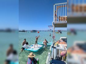 A lively scene at a sandbar with family and friends enjoying water activities, including a child shooting hoops on a floating mat — perfect for summer fun in the sun.