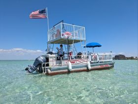 A vibrant pontoon boat with an upper deck and American flag, anchored in clear waters, showcasing a perfect spot for relaxation and fun on the water.