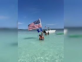 A lively group enjoys a sunny day in shallow waters, celebrating with flags and a boat in the background — perfect for summer fun and gatherings.