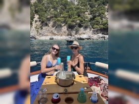 Two women toast with cocktails on a boat, enjoying a sunny day surrounded by stunning coastal scenery — a perfect moment for summer celebrations.
