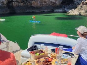 Guests enjoy a picturesque day on the water with paddleboarding and a delicious spread of snacks aboard their boat, surrounded by stunning green waters and rocky cliffs.