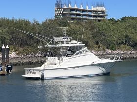 A sleek yacht named 'Fish Machine' is docked in calm waters, showcasing its elegant design against a backdrop of lush greenery and construction.