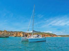 A lively group enjoys a day on a catamaran, sailing through beautiful turquoise waters with stunning coastal cliffs in the background — perfect for adventure seekers and relaxation alike.