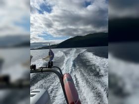A scenic view from the bow of a boat cutting through the water, showcasing the beautiful coastline and lush green hills in the background.