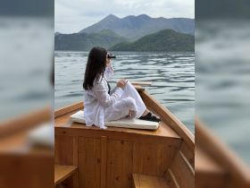 A woman enjoys a serene moment on a boat, using binoculars to take in the stunning mountain scenery — a perfect escape for nature lovers.