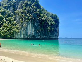 A stunning view of a beach with crystal-clear waters and a dramatic cliff backdrop, where people are enjoying swimming and relaxing — a perfect tropical getaway.