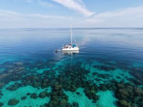 A stunning aerial view of a sailboat navigating through crystal-clear waters, showcasing vibrant coral formations beneath the surface — perfect for sailing enthusiasts and nature lovers.