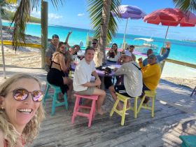 A joyful group of friends and family gather at a beachside table, enjoying drinks and the stunning turquoise waters — perfect for memorable celebrations in paradise.