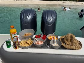 A vibrant spread of snacks and drinks is laid out on a boat, perfect for enjoying a day at the beach, with guests visible in the background enjoying the water.