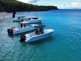 A lively scene with multiple boats anchored near a beach, where guests are enjoying swimming and socializing in the clear turquoise waters — a perfect day for aquatic adventures.