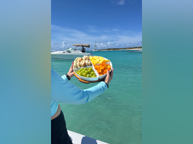 A vibrant platter of snacks, including grapes, mango, and chips, is held up against a stunning turquoise backdrop, perfect for enjoying on a boat trip.