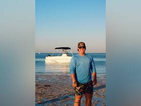 A man stands on the beach with a boat in the background, enjoying a sunny day by the water — perfect for beach outings and boating adventures.