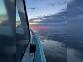 A serene view from the side of a boat as it glides over calm waters at sunset, showcasing the beautiful reflection of the sky.