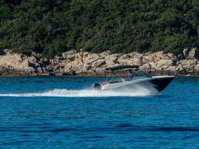 A sleek speedboat glides through calm waters, showcasing its design against a stunning natural backdrop of rocky shores and greenery.
