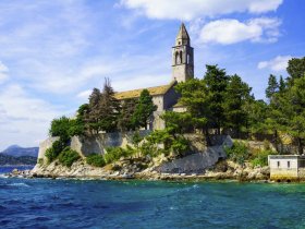 A picturesque view of a historic church perched on a rocky coastline, surrounded by lush greenery and clear blue waters — a stunning landmark perfect for sightseeing during boat tours.