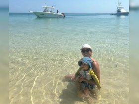 A joyful moment captured as a mother and her child enjoy the shallow waters at the beach, with boats anchored in the background — perfect for family outings and beach adventures.