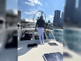 A man stands confidently on the deck of a boat with a stunning city skyline and ferris wheel in the background, capturing the essence of a vibrant day on the water.
