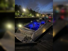 A sleek boat named 'Hey Buddy' is docked at night, illuminated by blue lights, with a vibrant city skyline in the background, perfect for evening cruises.