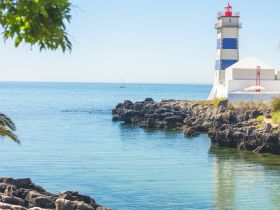 A picturesque view featuring a lighthouse by the coastline, with a sailboat in the distance and swimmers enjoying the calm waters — a perfect representation of a serene day by the sea.