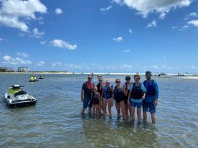 A cheerful group of friends and family enjoy a day at the beach, posing in shallow water with jet skis in the background — perfect for summer adventures and water sports.