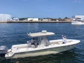 A fishing boat cruising through the water with two people onboard, one waving at the camera — a perfect snapshot of a day on the water.