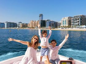 A joyful family enjoys a sunny day on a boat, waving with excitement against a stunning city backdrop — perfect for memorable outings and family adventures on the water.