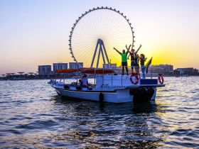 A lively group celebrates aboard a boat during sunset, with a stunning ferris wheel in the background — perfect for memorable boat tours and group outings.