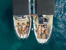 A fun group of friends relaxes on the deck of two boats, enjoying the sun and clear waters — perfect for a summer getaway or beach party.