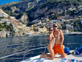 A couple poses on the deck of a boat with the stunning Amalfi Coast in the background, capturing a perfect moment of romance and adventure on the water.