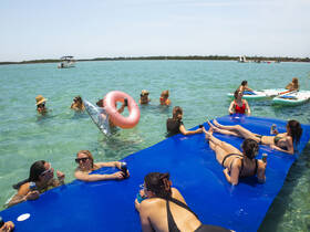 A lively group of friends enjoys a fun day in the water, lounging on a floating mat and sipping drinks, perfect for summer parties and beach outings.