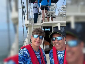 A father and son pose for a selfie on a boat, both wearing life jackets and vibrant shirts, capturing a memorable sailing experience together.