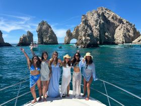 A lively group of friends celebrates on the deck of a boat in Cabo San Lucas, with stunning rock formations in the background — perfect for unforgettable summer adventures.