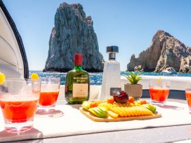 A vibrant display of cocktails and fresh fruit snacks on a boat, set against a stunning coastal backdrop — perfect for enjoying a luxurious day on the water.