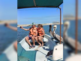 A couple takes a selfie on a small boat, enjoying a sunny day on the water with a pelican nearby — a perfect moment for wildlife enthusiasts.