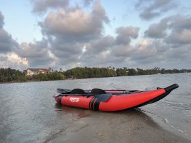 A vibrant red inflatable kayak rests on the sandy shore, surrounded by lush greenery and a scenic backdrop, perfect for exploring calm waters.