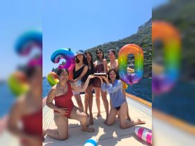 A joyful group of friends celebrates a birthday on a boat, showcasing colorful balloons and a cake against a stunning coastal backdrop — perfect for summer festivities.
