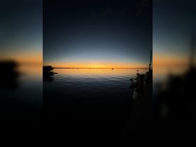 A serene sunset view over calm waters, with silhouettes of boats docked at the pier, creating a picturesque scene perfect for evening relaxation.