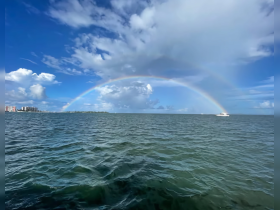 A stunning view of a rainbow arching over the ocean, with vibrant clouds and a distant boat, capturing the beauty of a perfect day on the water.