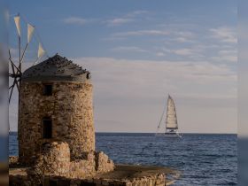 A picturesque view of a sailing boat gliding past a historic windmill along the coastline, capturing the essence of maritime beauty and local landmarks.