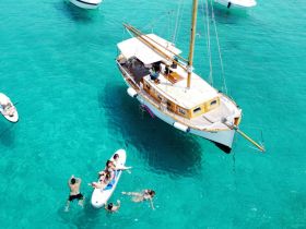 A lively scene featuring a group enjoying the turquoise waters, with some swimming and others on a paddleboard near a beautiful boat — perfect for summer adventures.