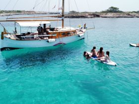 A lively group enjoys paddleboarding and swimming in crystal-clear turquoise waters near a charming boat, perfect for summer adventures and fun on the water.