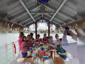 A lively group enjoys a celebration aboard a boat, raising their drinks in a toast while surrounded by delicious snacks — perfect for a fun day on the water.