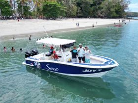A lively group poses on their boat near a beautiful beach, with people enjoying the water in the background — a perfect snapshot of a fun day on the water.