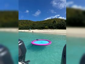 A vibrant floating mat is anchored in clear turquoise waters, with beachgoers enjoying the sun and sand in the background — perfect for a day of relaxation and fun on the water.