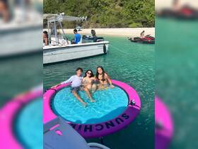 A lively group enjoys a sunny day on a floating mat in crystal-clear waters, surrounded by boats and a beautiful beach backdrop — perfect for summer fun and relaxation.